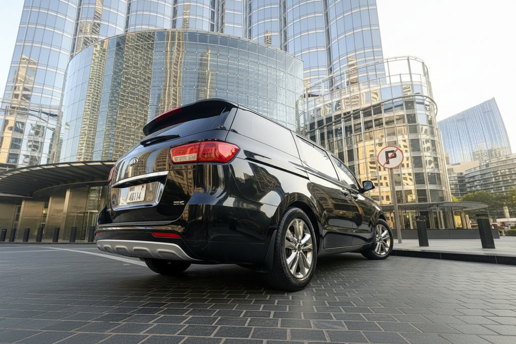 A black Kia Carnival minivan parked outdoors in front of a large modern glass skyscraper, viewed from a low angle.