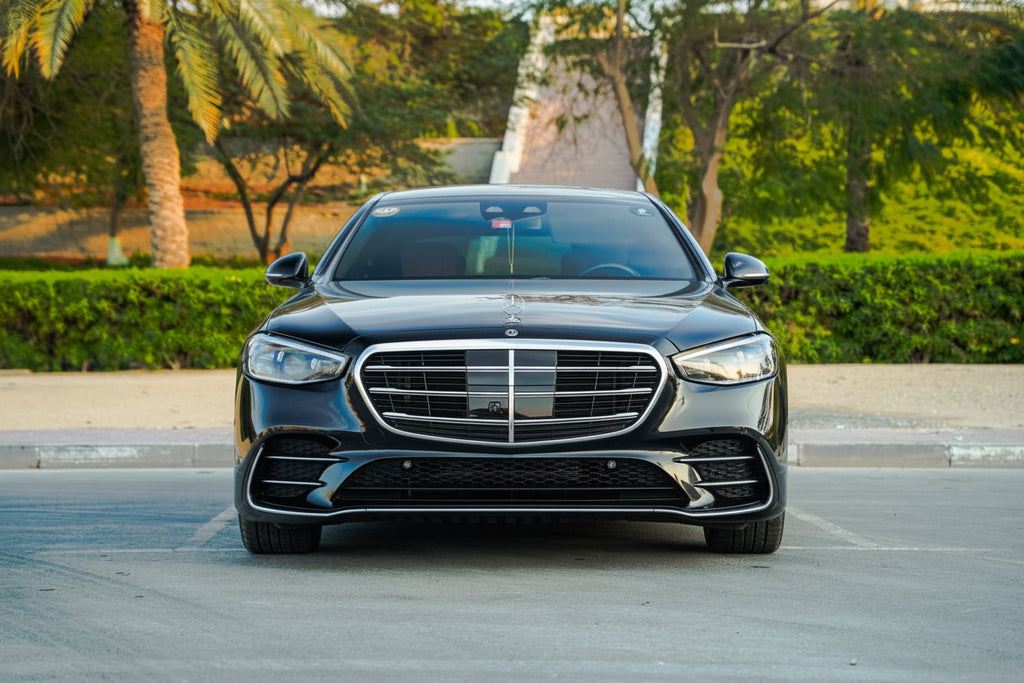 A black Mercedes-Benz S-Class sedan photographed from the front in an outdoor parking area with trees and hedges in the background.