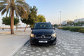 A black Mercedes-Benz minivan parked on a paved street outdoors, with palm trees, buildings, and a clear sky in the background.